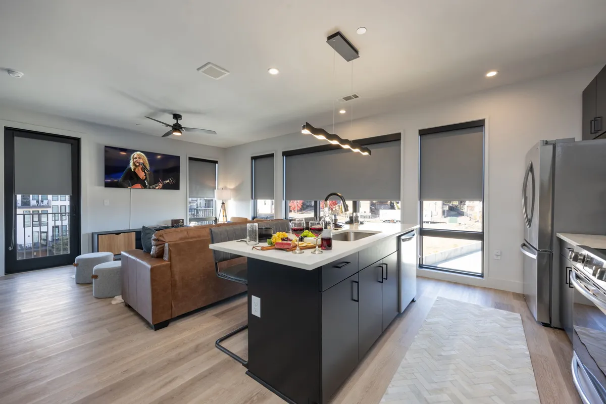 An open-concept interior featuring a modern kitchen island with seating, a comfortable sofa, a large television, and expansive floor-to-ceiling windows that bathe the space in natural light. The room seamlessly integrates the kitchen, living, and dining areas, emphasizing communal living and entertaining.
