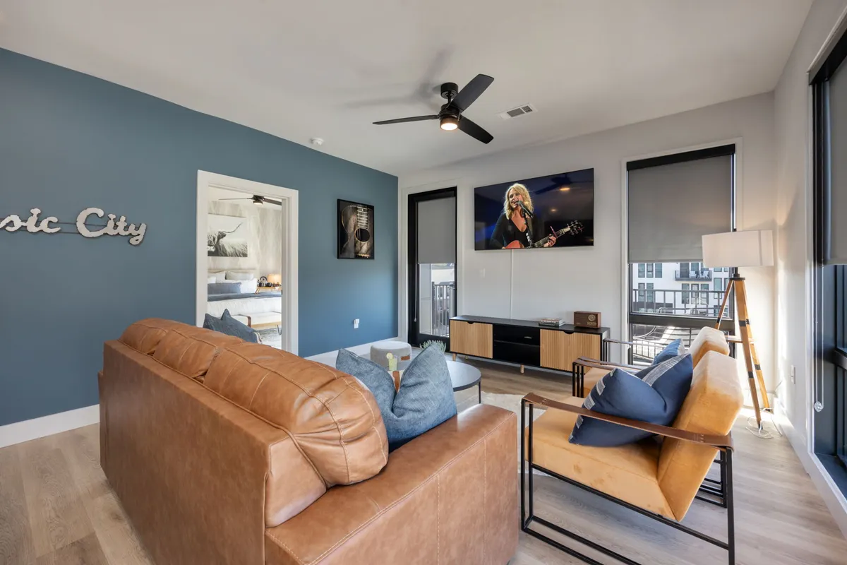 A cozy, modern living room with a leather sofa, a television displaying a musician in action, decorative wall art, and expansive windows letting in natural light. The space feels intimate yet lively, suitable for movie nights or casual entertaining.