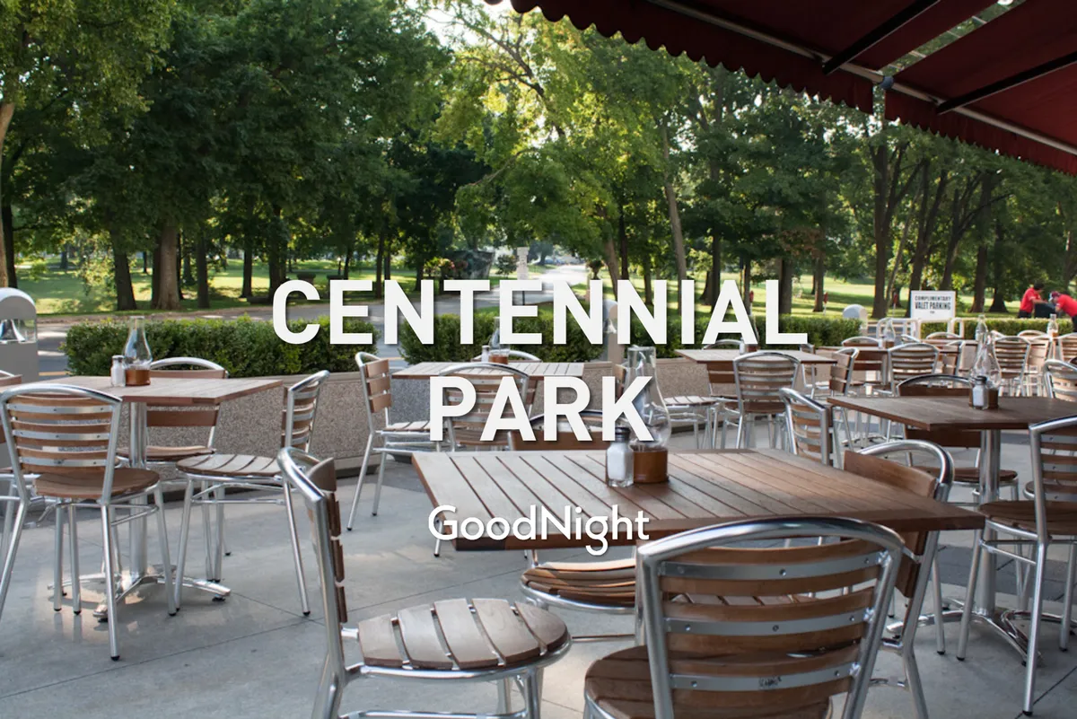 A spacious outdoor seating area shaded by a canopy and surrounded by lush green trees. Wooden tables are arranged for group dining or relaxed gatherings, inviting guests to enjoy meals or socialize in comfort. The space is infused with natural light and a casual, resort-like atmosphere.