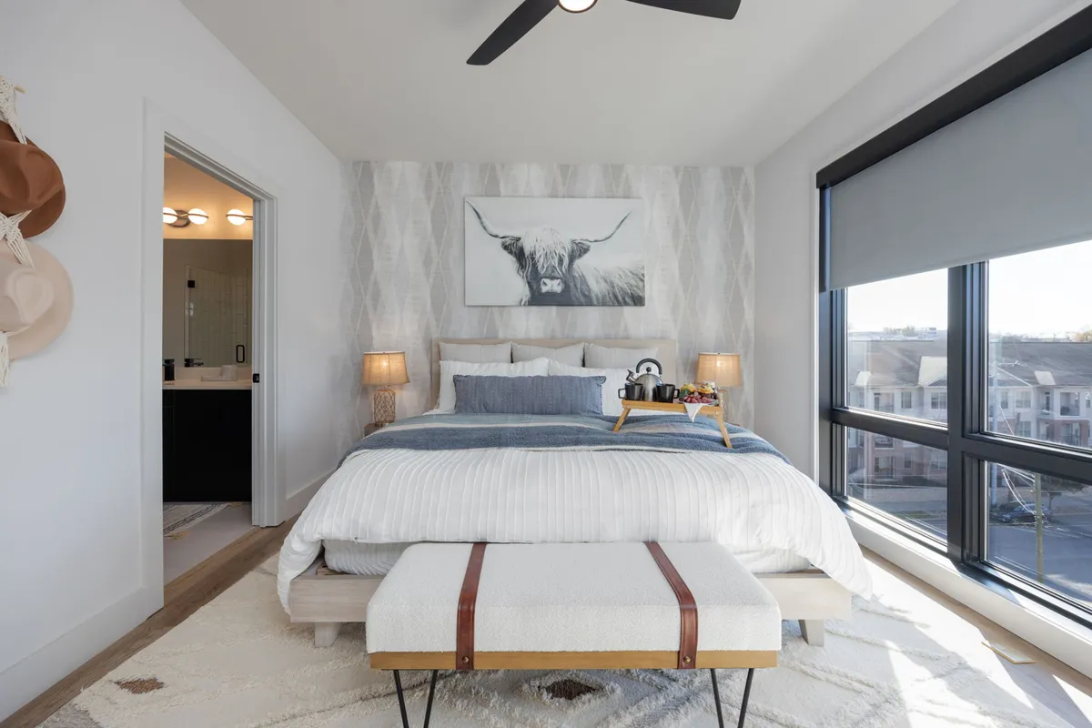 A tranquil primary bedroom featuring a large bed with crisp white and blue bedding, framed artwork of a cow gracefully displayed above the headboard, stylish nightstands with modern lamps, a plush ottoman at the foot of the bed, and large windows that fill the room with natural light.
