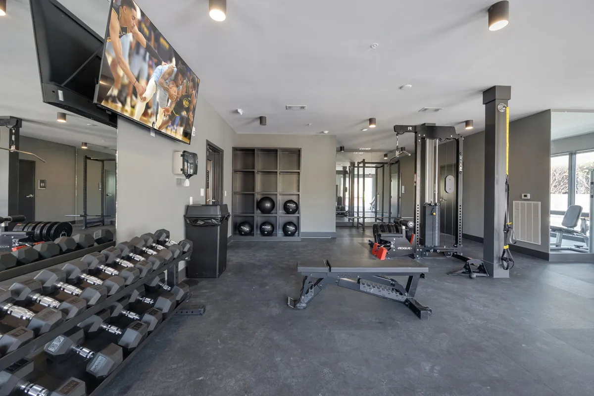 A modern gym space with a dumbbell rack, weight bench, TV screen, and various exercise equipment. The room is bright and energetic, inviting residents to maintain fitness routines or join group classes in a contemporary setting.