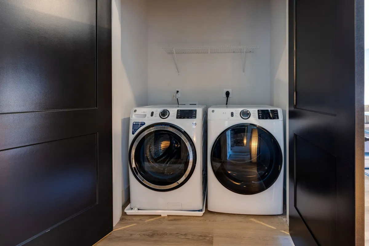 The image displays an in-unit laundry area featuring a washer, dryer, and a practical shelf for storing laundry and cleaning supplies. The clean setup suggests ease of access and efficient organization, highlighting modern conveniences within the unit.